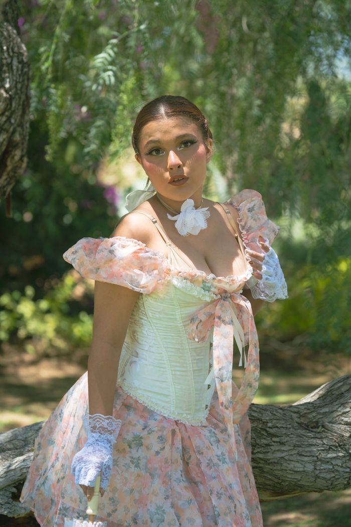 who-we-are A woman in a vintage floral dress poses elegantly in a sunlit garden, showcasing timeless beauty and style.