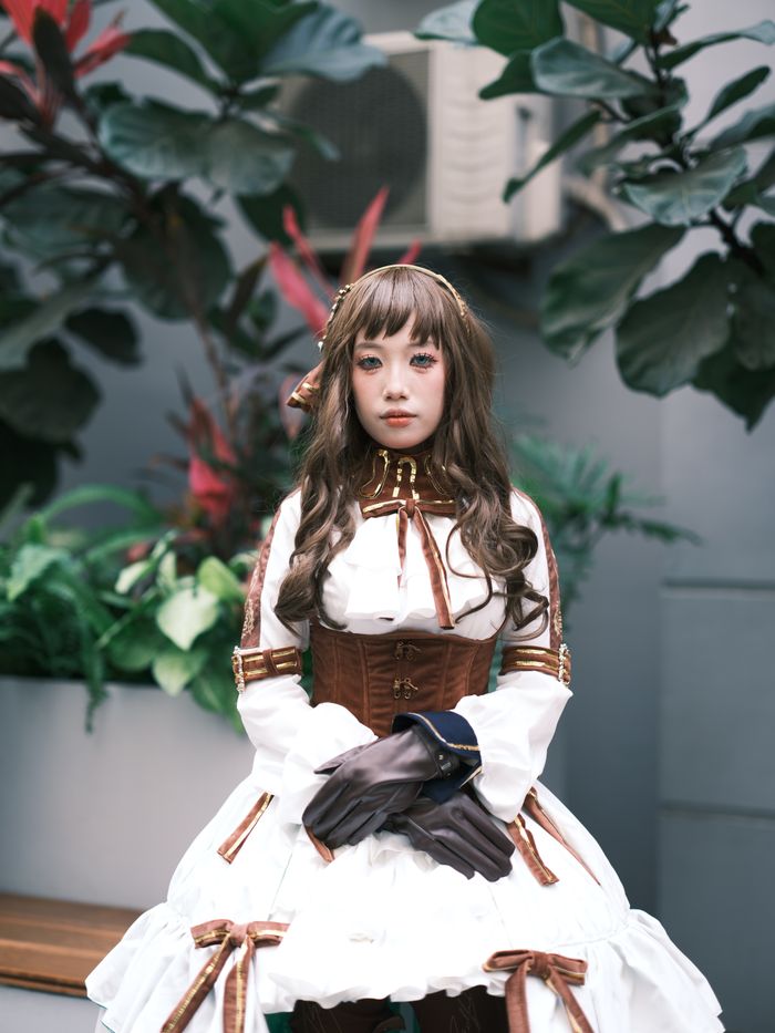 services-01 A cosplay model poses in intricate vintage fashion with rich brown and white tones, surrounded by lush foliage.